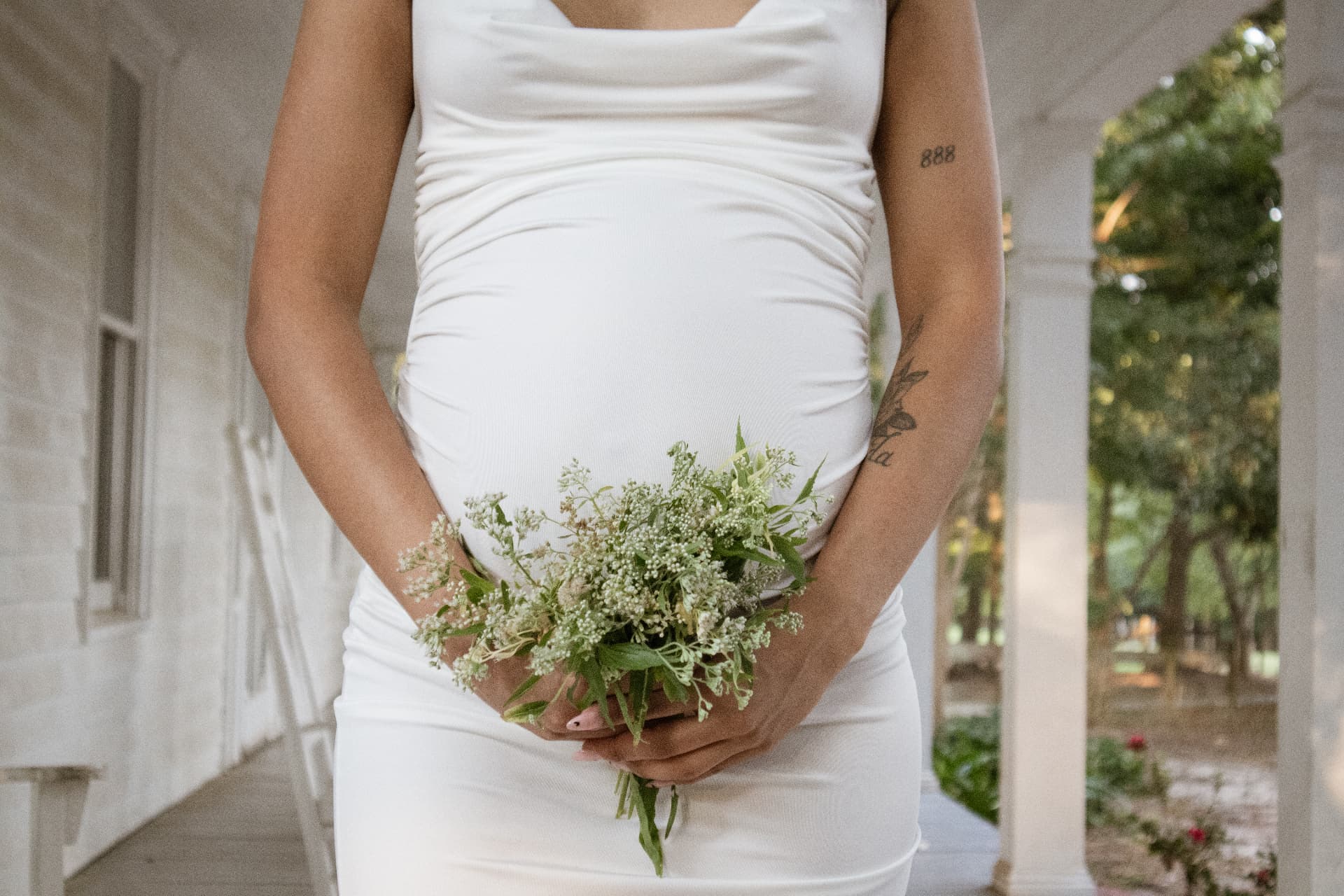 Expecting mother in a white dress cradling her bump and holding wildflowers on a porch